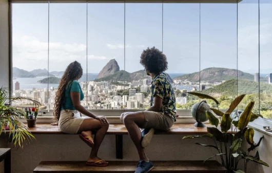 Imagem de duas pessoas sentadas na bancada de um apartamento olhando o Pão de Açúcar, no Rio de Janeiro, pela janela para ilustrar matéria sobre as cidades mais visitadas do Brasil