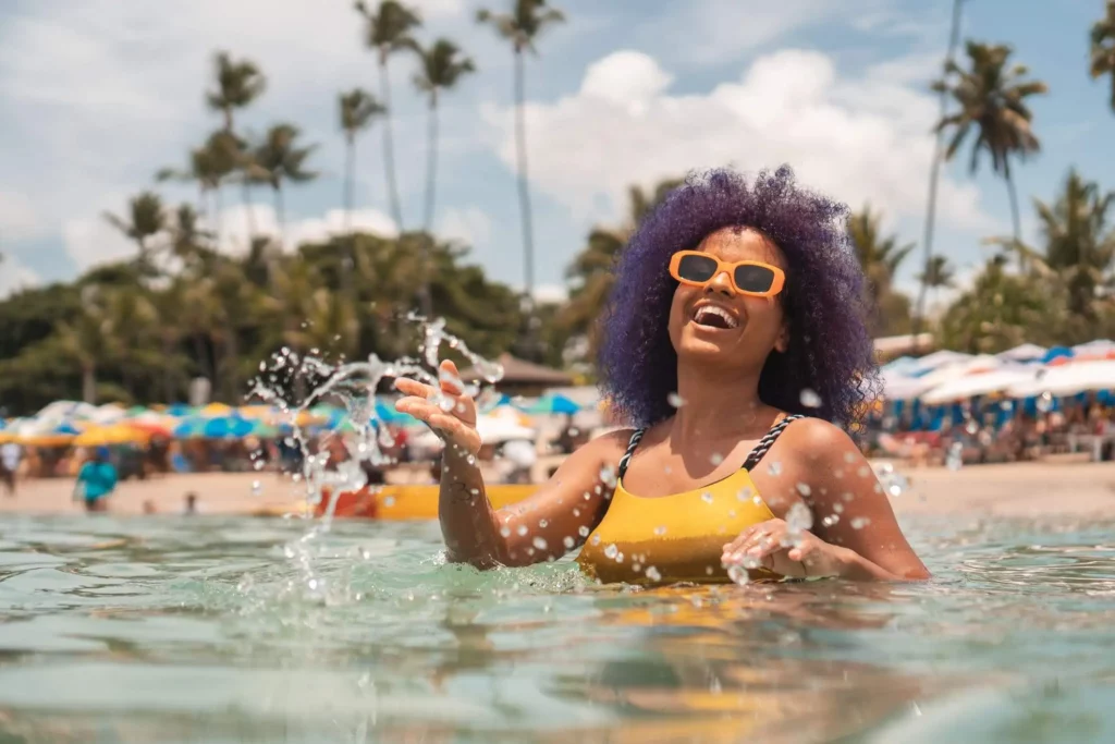 Imagem de uma mulher negra com óculos de sol amarelo dentro do mar sorrindo para ilustrar matéria sobre as cidades mais visitadas no verão em todo o Brasil
