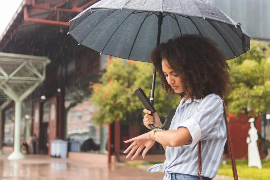 Imagem de uma mulher negra embaixo de um guarda chuva olhando para o relógio no pulso para ilustrar matéria sobre as cidades que mais chovem no Brasil