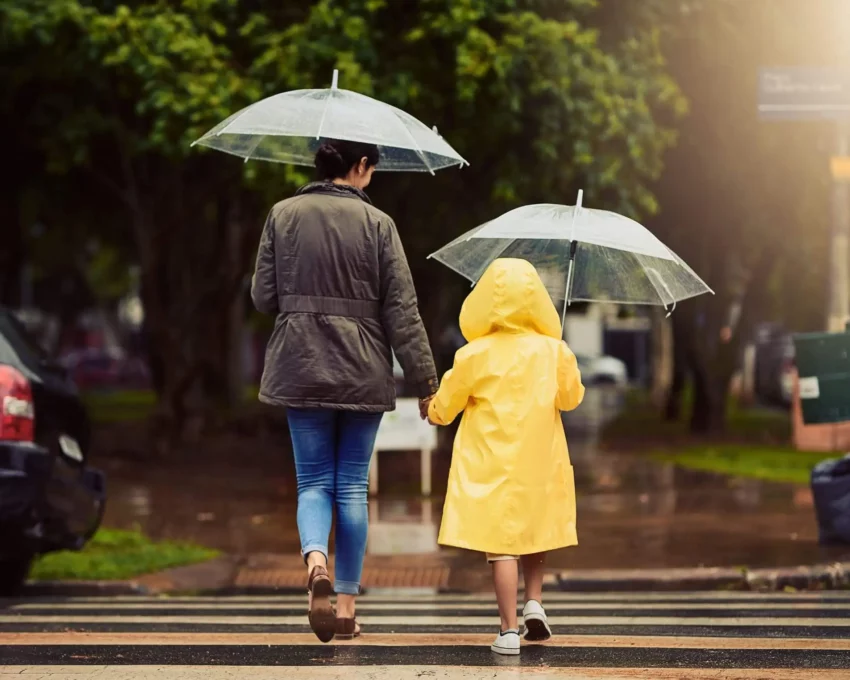 Imagem de uma mulher e uma criança de costas andando em uma faixa de pedestres com guarda chuva na mão para ilustrar matéria sobre as cidades que mais chovem no Brasil