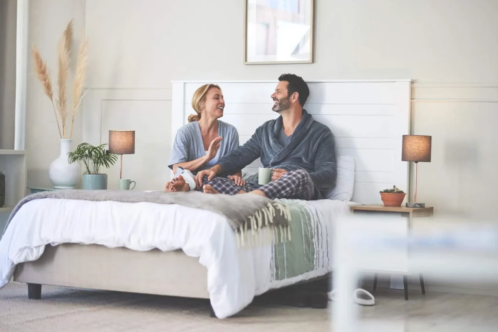 Imagem de uma mulher e um homem tomando café na cama de casal em um quarto claro com decorações minimalistas para ilustrar matéria sobre cores para pintar quarto de casal
