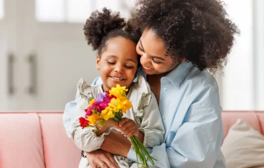 Foto que ilustra matéria sobre decoração para dia das mães mostra uma menina sentada no colo de sua mãe enquantro segura um buquê de flores (Foto: Shutterstock)