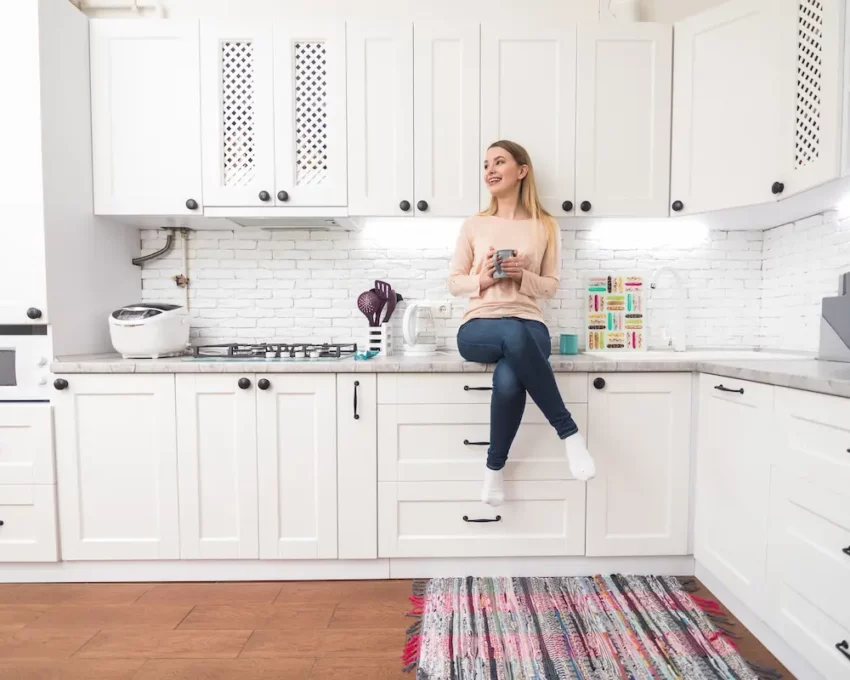 Cozinha planejada: imagem de uma mulher sentada na bancada de uma cozinha iluminada com armários brancos e piso de madeira