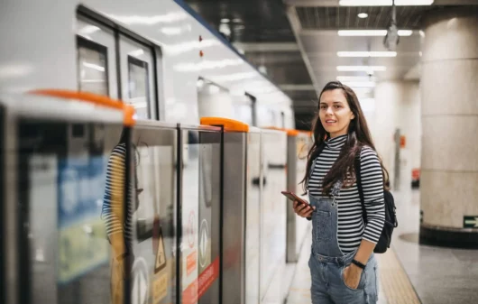 Imagem de uma mulher jovem sorrindo com o celular na mão enquanto aguarda o trem em uma estação de metrô para ilustrar matéria sobre Linha 9-Esmeralda